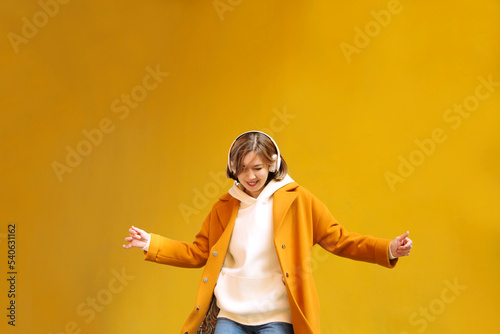 a woman in a yellow coat with headphones dances and sings on the street of the city against the background of a yellow wall