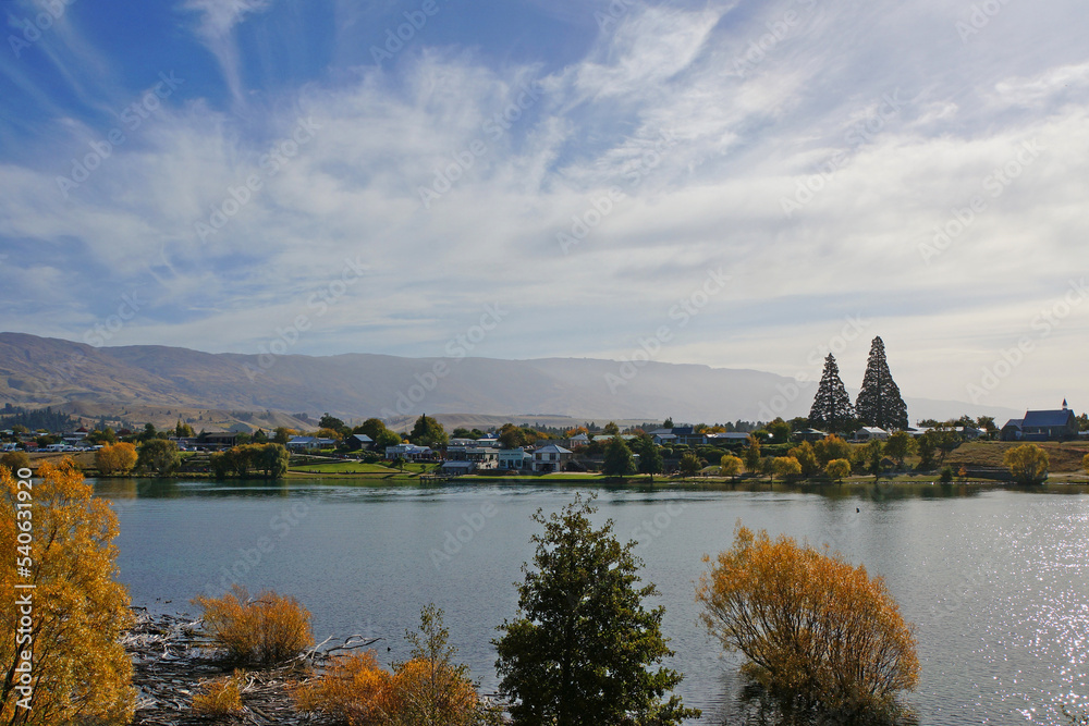 Obraz premium A view across Lake Dunstan to old Cromwell town from Cornish Point on the Lake Dunstan Cycle Trail. A sunny Autumn day under a blue sky with the Piza mountain range in the distance.