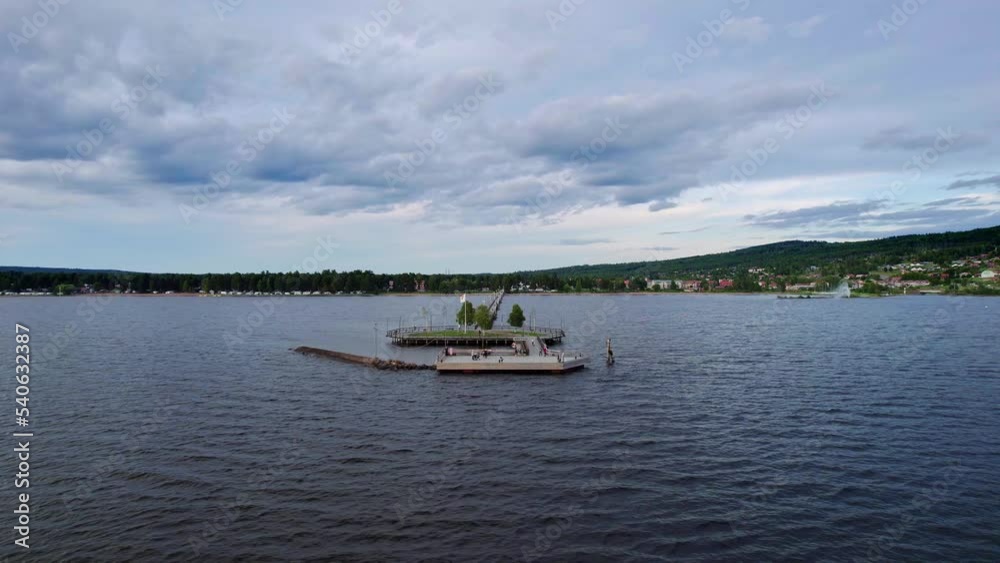 Arc shot: A wooden jetty with tourists enjoying the view over the lake and town of Rattvik. At the pier a peninsula with wooden promenade and trees. In Dalarna Sweden, Summer Season. Arc shot: A wooden jetty with tourists enjoying the view over the lake and town of Rattvik. At the pier a peninsula with wooden promenade and trees. In Dalarna Sweden, Summer Season.