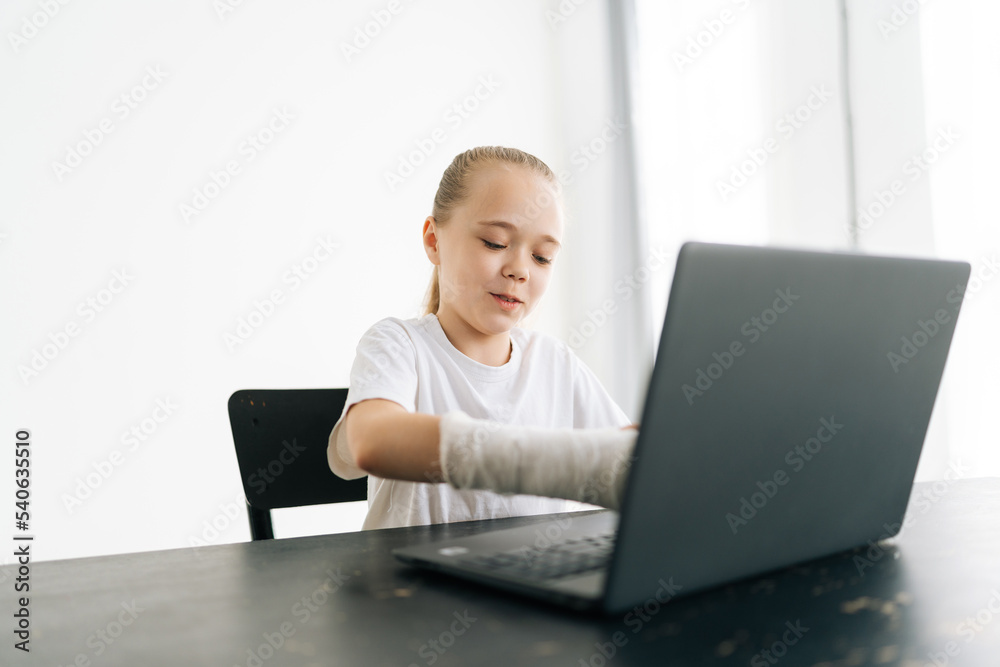 Portrait of cute little girl with broken hand wrapped in white plaster bandage typing on laptop computer looking to screen sitting at table in light room. Concept of child insurance and healthcare. Portrait of cute little girl with broken hand wrapped in white plaster bandage typing on laptop computer looking to screen sitting at table in light room. Concept of child insurance and healthcare.