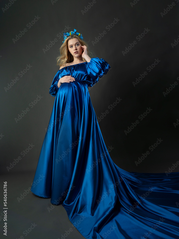 A pregnant girl with blonde hair in a long blue dress and a headband with butterflies. A full-length photo on a dark background. The concept of a happy pregnancy.