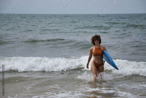 Attractive mature woman with curly hair, sunglasses and bikini, coming out of the water holding a blue surfboard under her arm. Concept sea, sand, sun, beach, vacation, surf, summer.