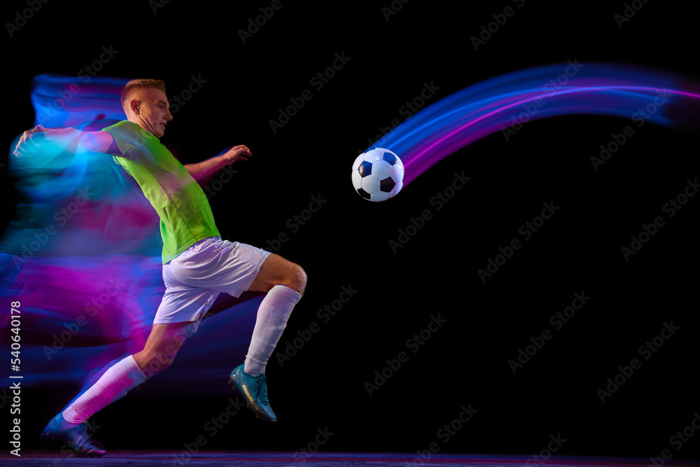 Sport in action. One man professional soccer player training with football ball isolated on dark background in neon light filter. Sport, speed, power and energy Sport in action. One man professional soccer player training with football ball isolated on dark background in neon light filter. Sport, speed, power and energy