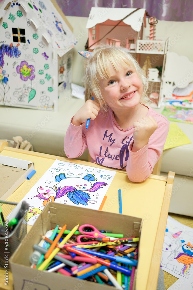 Fototapeta premium A blonde-haired child girl paints a drawing of a unicorn in a children's coloring book. A cheerful child is engaged in fine art.
