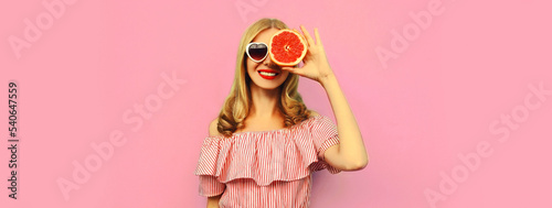 Summer portrait of happy smiling young woman with slice of juicy grapefruit wearing heart shaped sunglasses on pink background