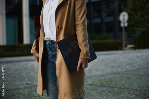 Millennial business woman wearing coat walking at city street with laptop at summer day.