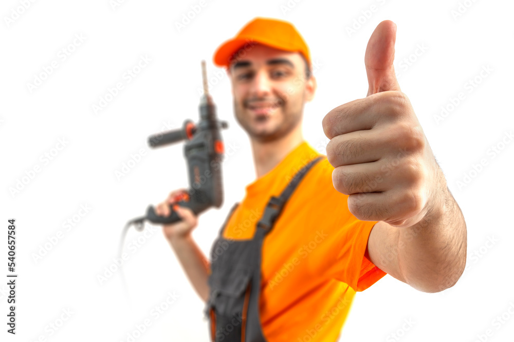 Young worker, thumb up, drilling with electric drill in his hands in blur. Craftsman repairman wearing orange uniform cap and t-shirt, overalls on a white isolated background for advertising.