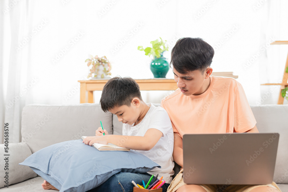 asian children and his father relax on living room, a boy learning to write and a man work from home, they sitting on sofa, happiness in family time asian children and his father relax on living room, a boy learning to write and a man work from home, they sitting on sofa, happiness in family time
