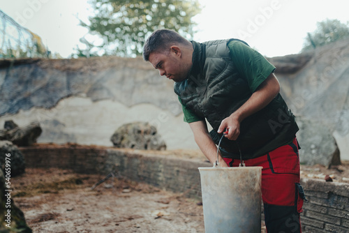 Caretaker with down syndrome in zoo giving food in animal enclosure. Concept of integration people with disabilities into society.