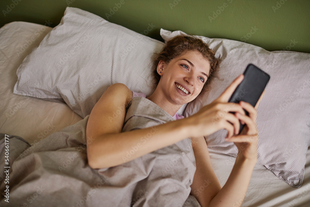 Young happy woman reading message in her smartphone and smiling while resting in her bed during weekends Young happy woman reading message in her smartphone and smiling while resting in her bed during weekends