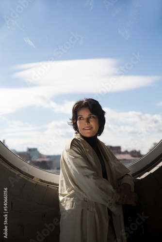 middle aged woman in a white cloak relaxing against the background of a round window with a blue sky and a view of the city. 