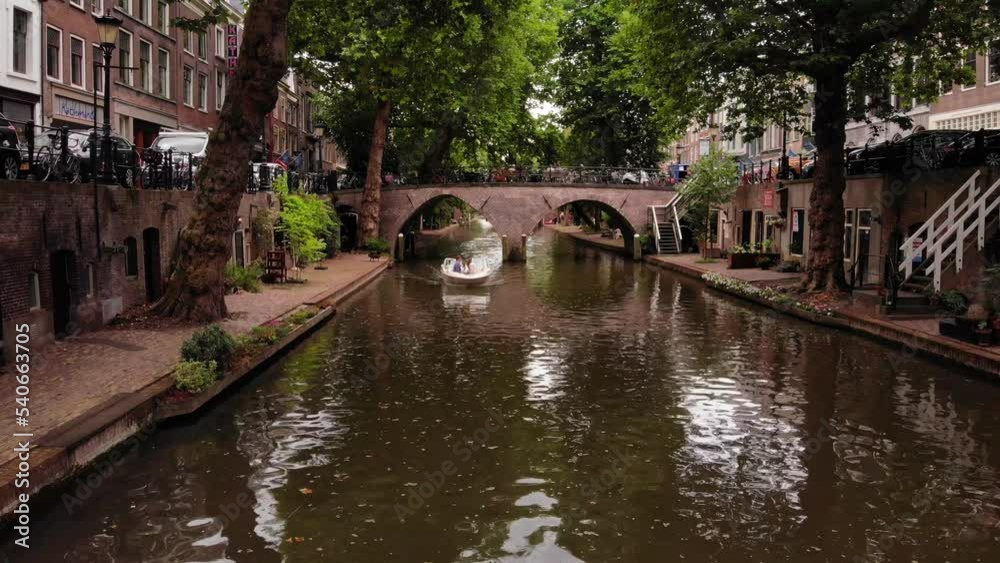 Motor boat passing under old bridge over Oudegracht water canal surrounded by grown trees on embankments in historic city centre. Utrecht, Netherlands