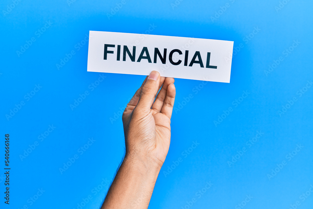 Hand of caucasian man holding paper with financial word over isolated blue background Hand of caucasian man holding paper with financial word over isolated blue background