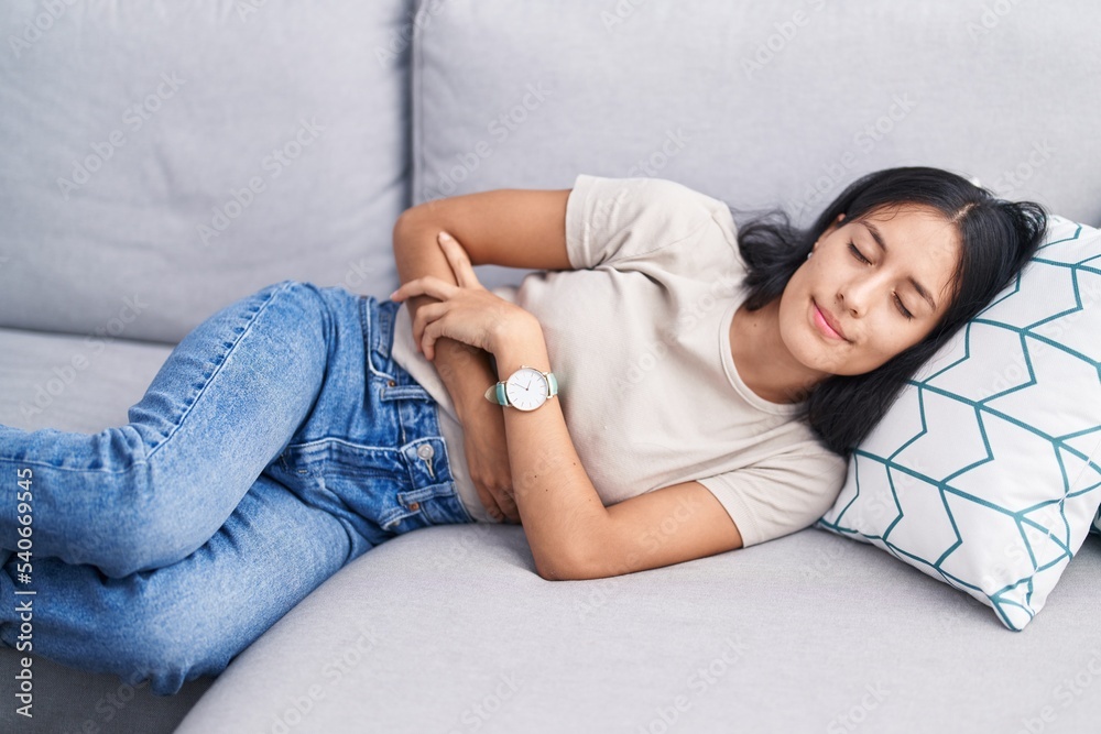 Young beautiful hispanic woman suffering for stomach ache lying on sofa at home Young beautiful hispanic woman suffering for stomach ache lying on sofa at home