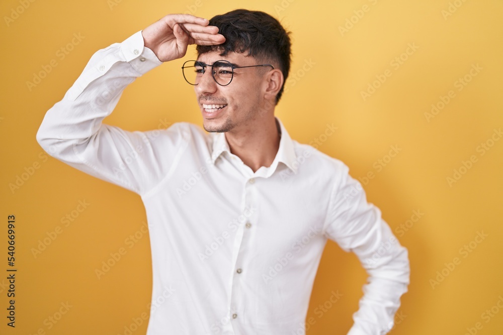 Young hispanic man standing over yellow background very happy and smiling looking far away with hand over head. searching concept. Young hispanic man standing over yellow background very happy and smiling looking far away with hand over head. searching concept.