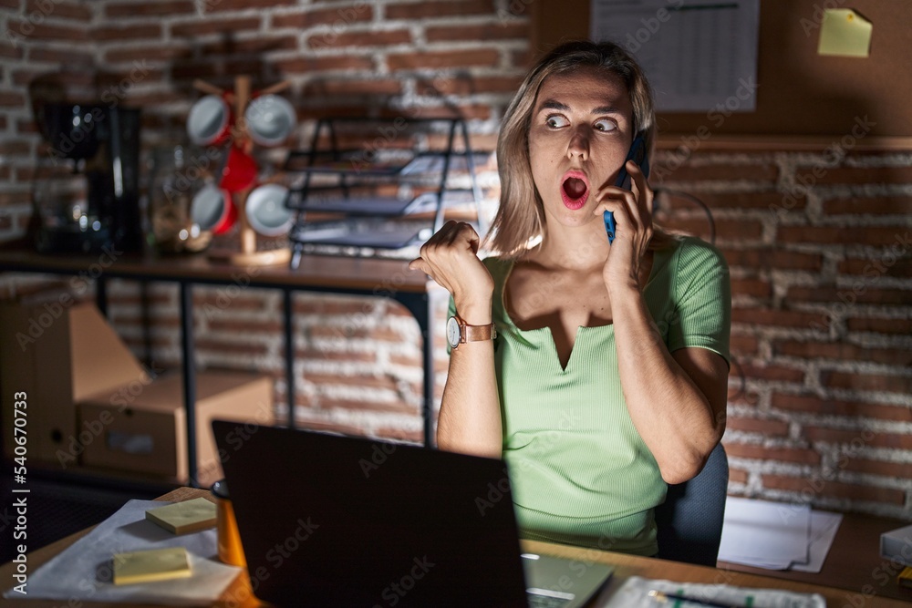 Young beautiful woman working at the office at night speaking on the phone surprised pointing with hand finger to the side, open mouth amazed expression. Young beautiful woman working at the office at night speaking on the phone surprised pointing with hand finger to the side, open mouth amazed expression.