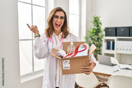 Young doctor woman holding box with medical items smiling happy pointing with hand and finger to the side