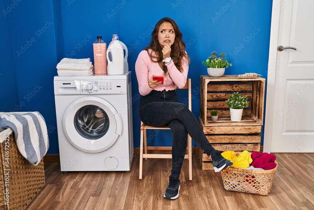 Young hispanic woman sitting waiting for laundry using smartphone thinking worried about a question, concerned and nervous with hand on chin Young hispanic woman sitting waiting for laundry using smartphone thinking worried about a question, concerned and nervous with hand on chin