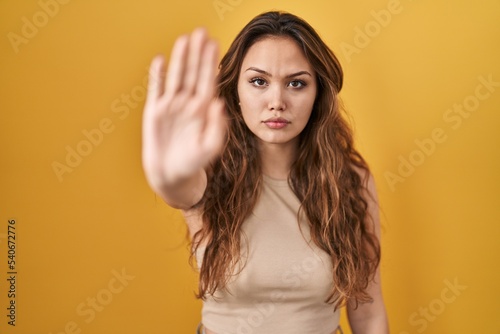 Young hispanic woman standing over yellow background doing stop sing with palm of the hand. warning expression with negative and serious gesture on the face.