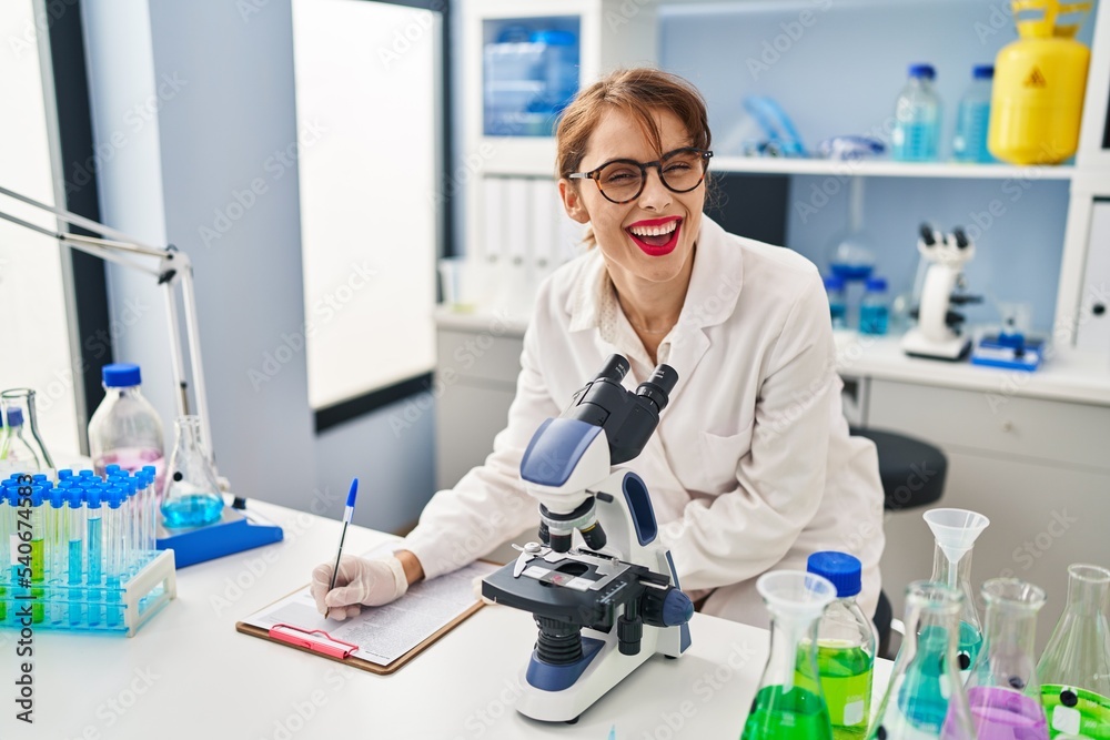 Young caucasian woman wearing scientist uniform writing on clipboard and uisng microscope at laboratory Young caucasian woman wearing scientist uniform writing on clipboard and uisng microscope at laboratory
