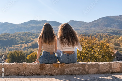 two girls are sitting with their backs against a beautiful landscape of autumn forest and mountains. Girls on the background of incredibly beautiful nature. Landscape of mountain ranges