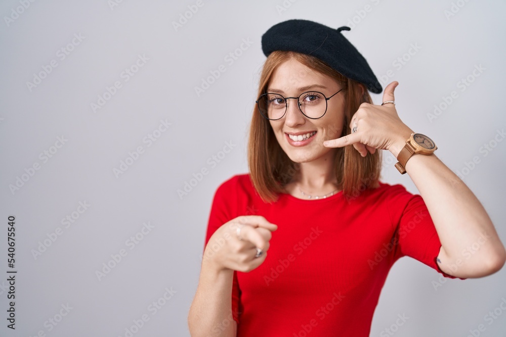 Young redhead woman standing wearing glasses and beret smiling doing talking on the telephone gesture and pointing to you. call me. Young redhead woman standing wearing glasses and beret smiling doing talking on the telephone gesture and pointing to you. call me.