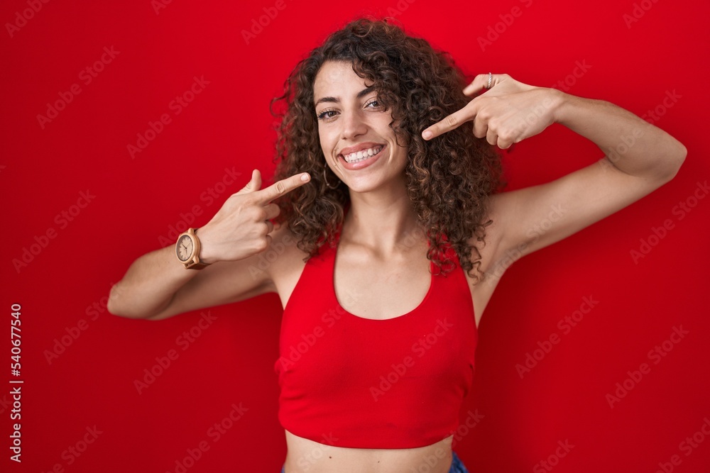 Hispanic woman with curly hair standing over red background smiling cheerful showing and pointing with fingers teeth and mouth. dental health concept.