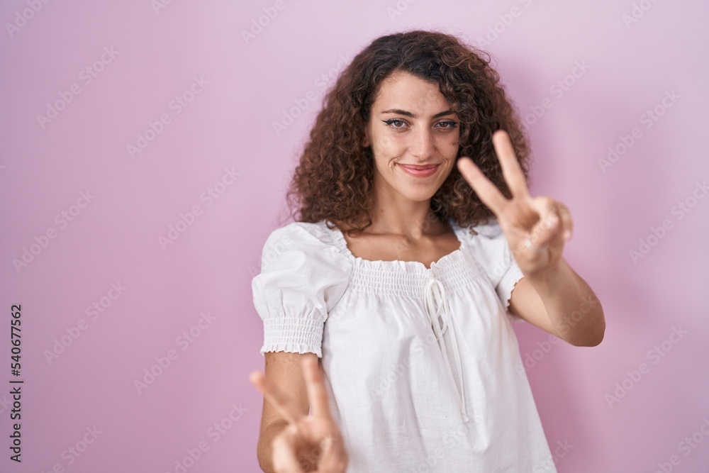 Hispanic woman with curly hair standing over pink background smiling looking to the camera showing fingers doing victory sign. number two. Hispanic woman with curly hair standing over pink background smiling looking to the camera showing fingers doing victory sign. number two.
