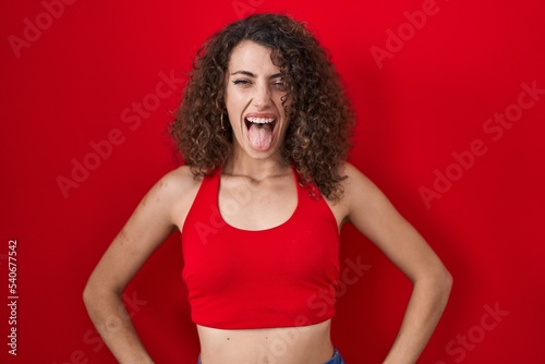 Hispanic woman with curly hair standing over red background sticking tongue out happy with funny expression. emotion concept.