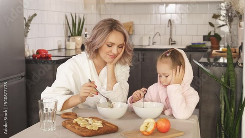 mom and little daughter sitting at the kitchen table do not want to eat oatmeal porridge with fruit, they are thinking about whether to eat porridge or not