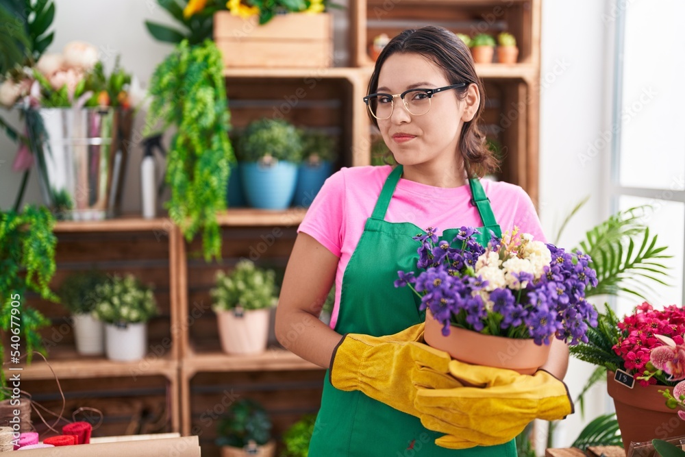 Hispanic young woman working at florist shop holding plant smiling looking to the side and staring away thinking.