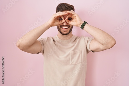 Hispanic man with beard standing over pink background doing ok gesture like binoculars sticking tongue out, eyes looking through fingers. crazy expression.
