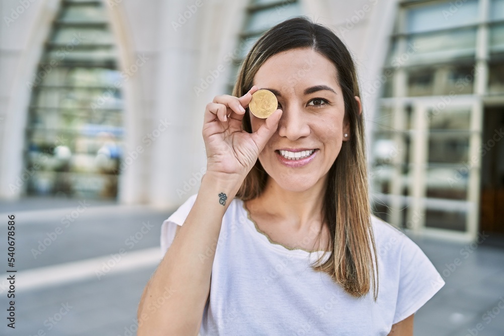 Young hispanic woman smiling confident holding bitcoin over eye at street