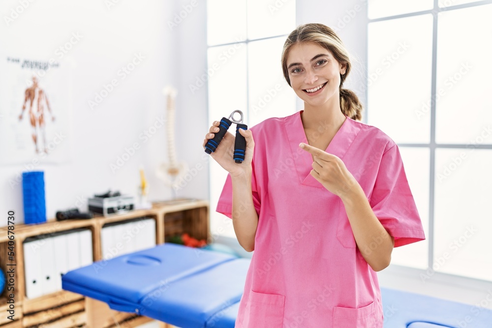 Young blonde woman working at pain recovery clinic holding hand strengthener smiling happy pointing with hand and finger