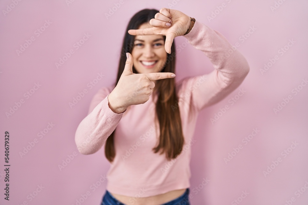 Young brunette woman standing over pink background smiling making frame with hands and fingers with happy face. creativity and photography concept. Young brunette woman standing over pink background smiling making frame with hands and fingers with happy face. creativity and photography concept.