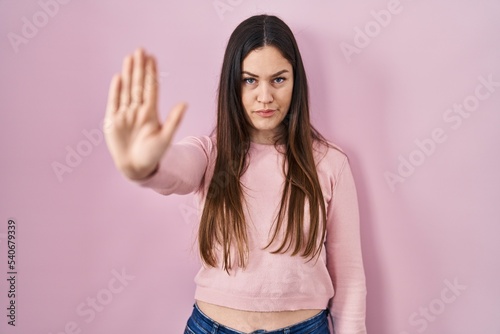 Young brunette woman standing over pink background doing stop sing with palm of the hand. warning expression with negative and serious gesture on the face.