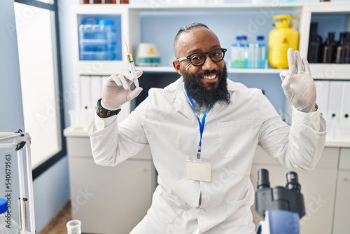 African american man working at scientist laboratory holding syringe doing ok sign with fingers, smiling friendly gesturing excellent symbol
