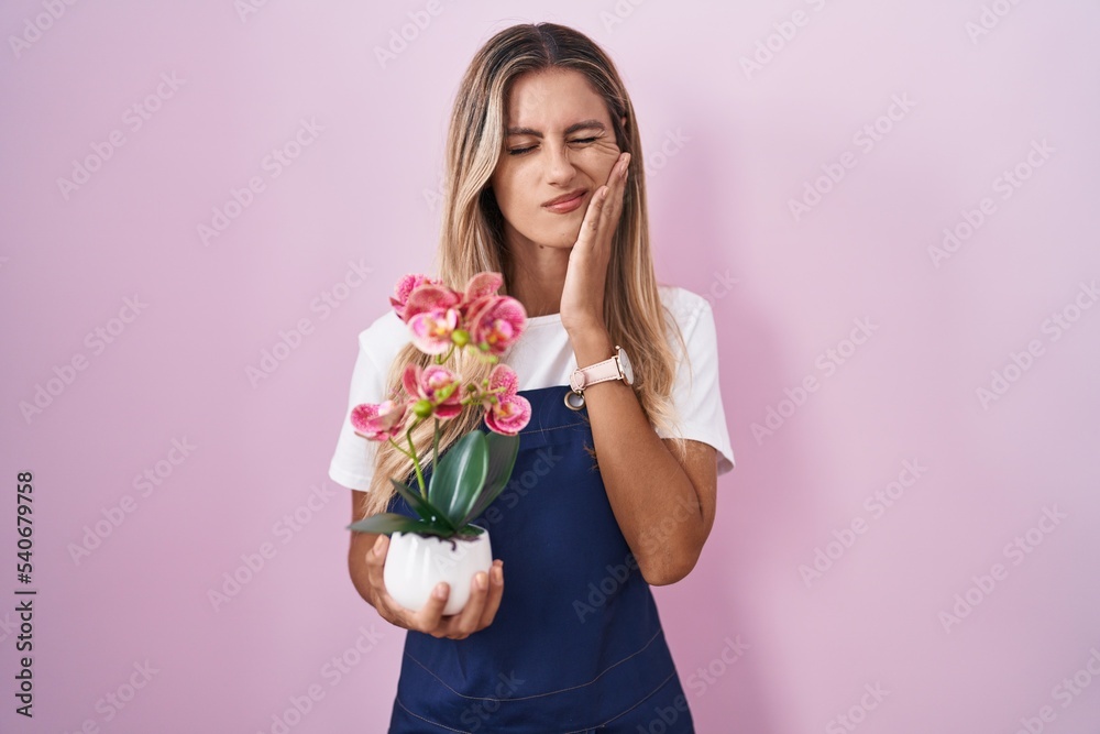 Young blonde woman wearing gardener apron holding plant touching mouth with hand with painful expression because of toothache or dental illness on teeth. dentist concept.