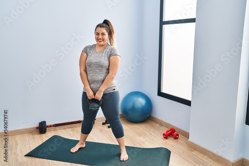 Young beautiful plus size woman smiling confident using kettlebell training at sport center