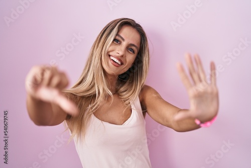 Young blonde woman standing over pink background showing and pointing up with fingers number six while smiling confident and happy.