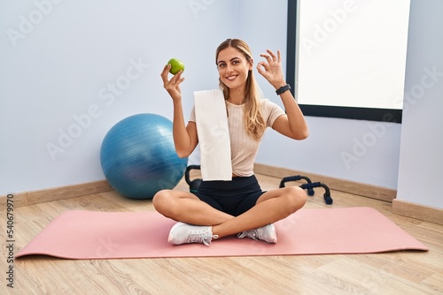 Young blonde woman wearing sportswear and towel holding healthy apple smiling positive doing ok sign with hand and fingers. successful expression.