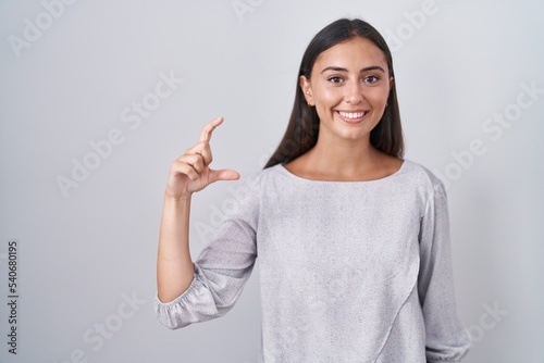 Young hispanic woman standing over white background smiling and confident gesturing with hand doing small size sign with fingers looking and the camera. measure concept.