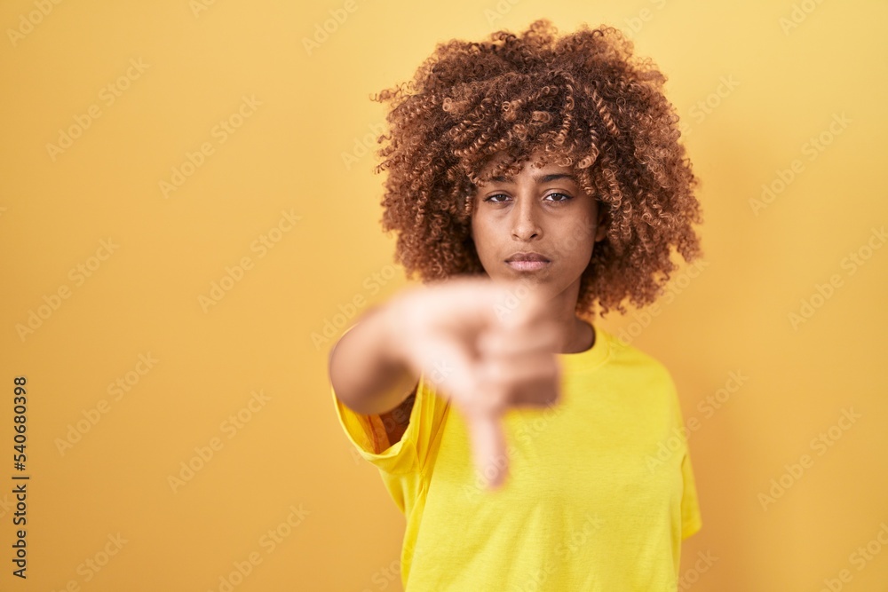 Young hispanic woman with curly hair standing over yellow background looking unhappy and angry showing rejection and negative with thumbs down gesture. bad expression. Young hispanic woman with curly hair standing over yellow background looking unhappy and angry showing rejection and negative with thumbs down gesture. bad expression.