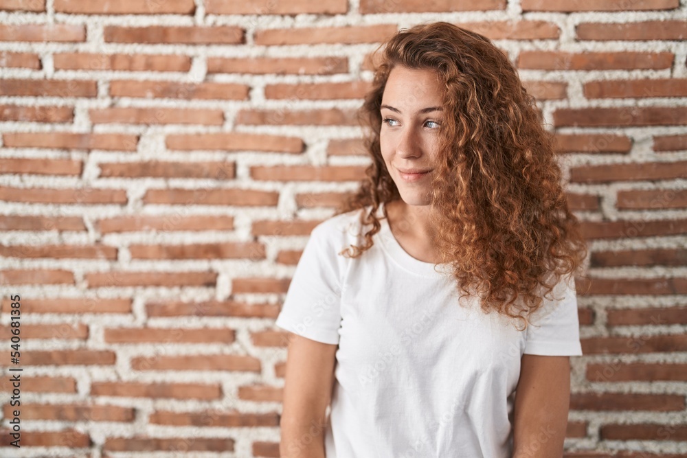 Young caucasian woman standing over bricks wall background looking away to side with smile on face, natural expression. laughing confident.