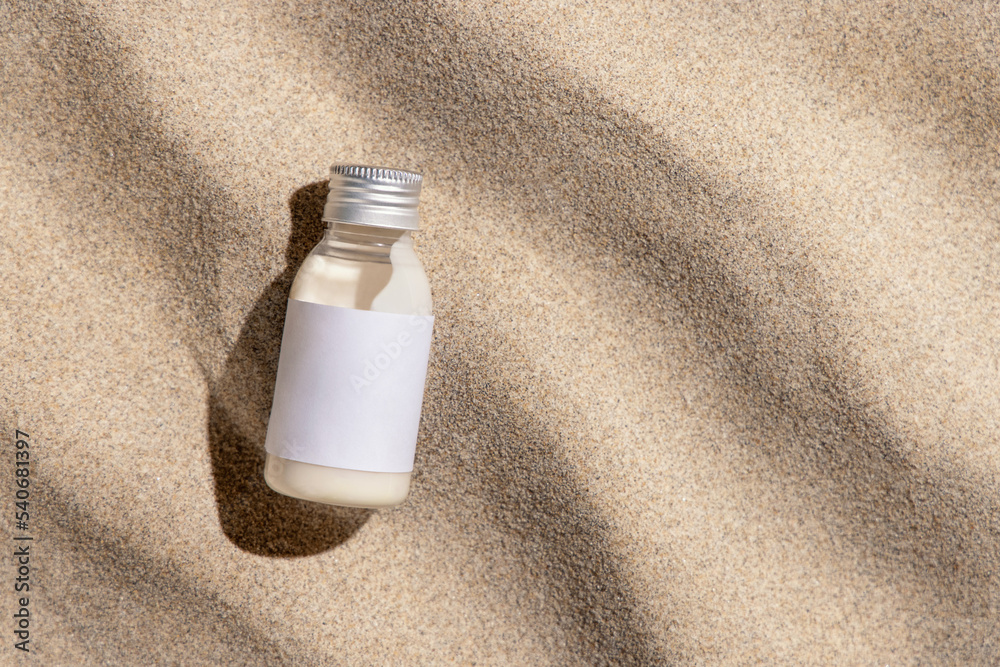 Cream bottle with blank label on beige sand top view, hard light. Cosmetic packaging mockup