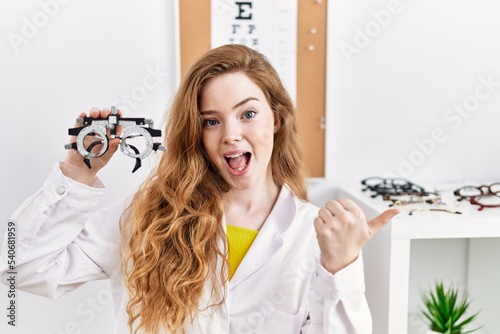 Young caucasian optician woman holding optometry glasses pointing thumb up to the side smiling happy with open mouth