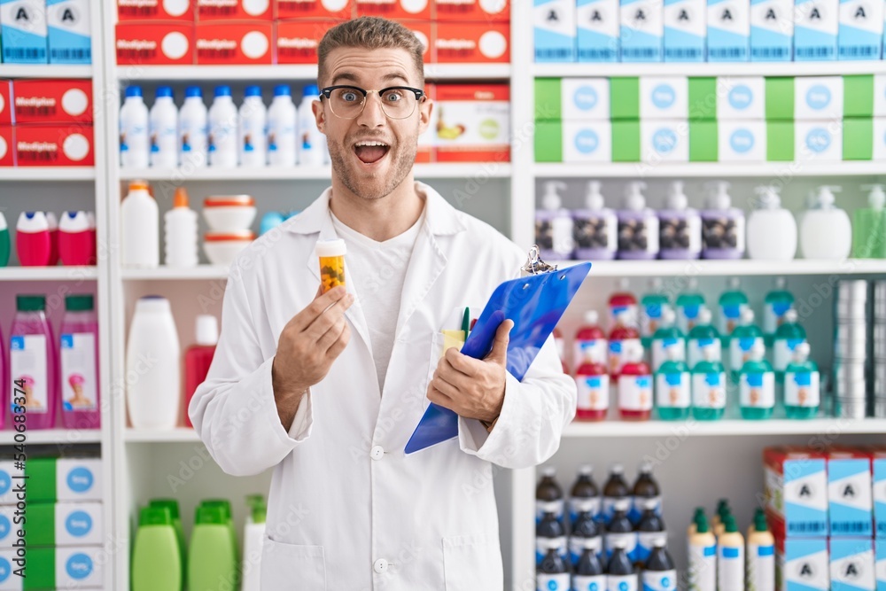 Young caucasian man working at pharmacy drugstore holding pills celebrating crazy and amazed for success with open eyes screaming excited. Young caucasian man working at pharmacy drugstore holding pills celebrating crazy and amazed for success with open eyes screaming excited.