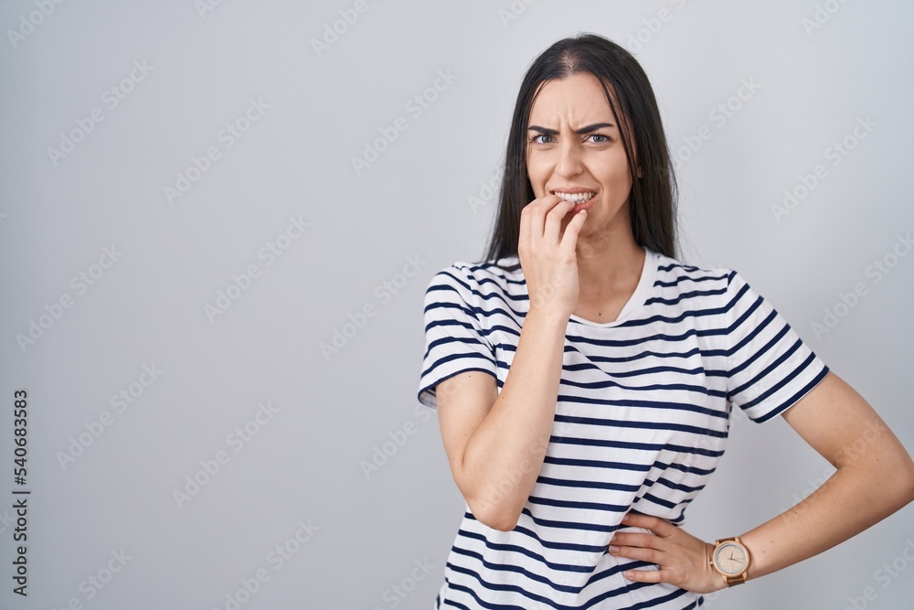 Young brunette woman wearing striped t shirt looking stressed and nervous with hands on mouth biting nails. anxiety problem. Young brunette woman wearing striped t shirt looking stressed and nervous with hands on mouth biting nails. anxiety problem.