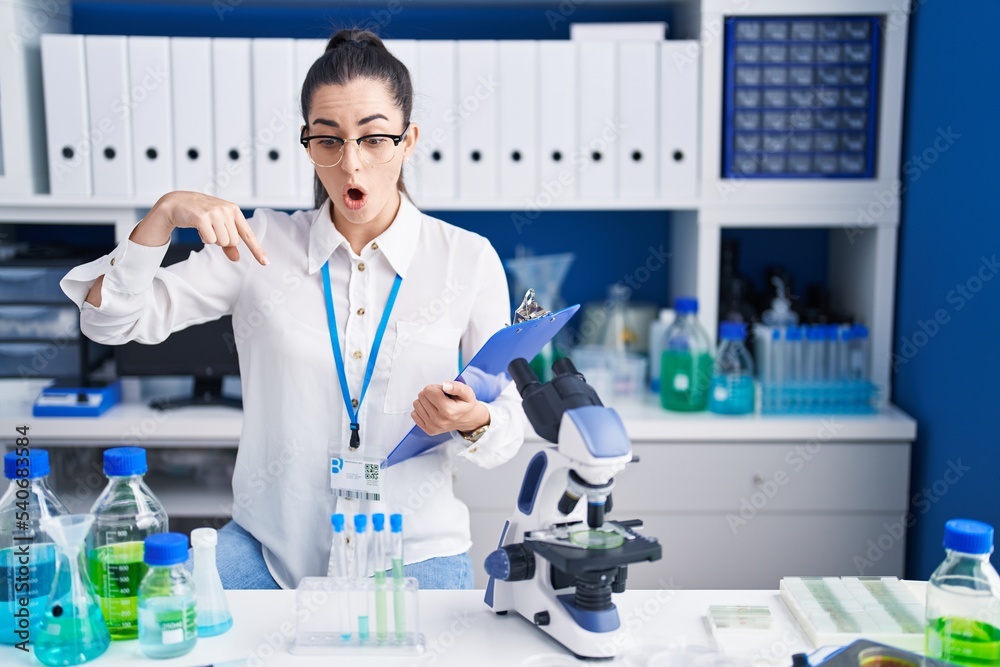 Young brunette woman working at scientist laboratory pointing down with fingers showing advertisement, surprised face and open mouth Young brunette woman working at scientist laboratory pointing down with fingers showing advertisement, surprised face and open mouth