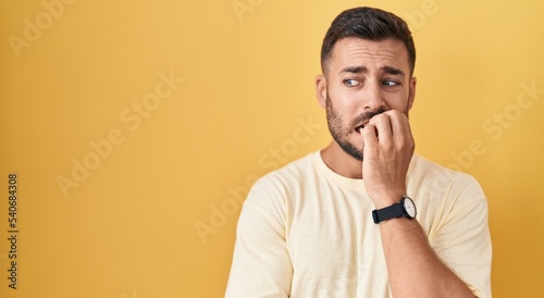 Handsome hispanic man standing over yellow background looking stressed and nervous with hands on mouth biting nails. anxiety problem.